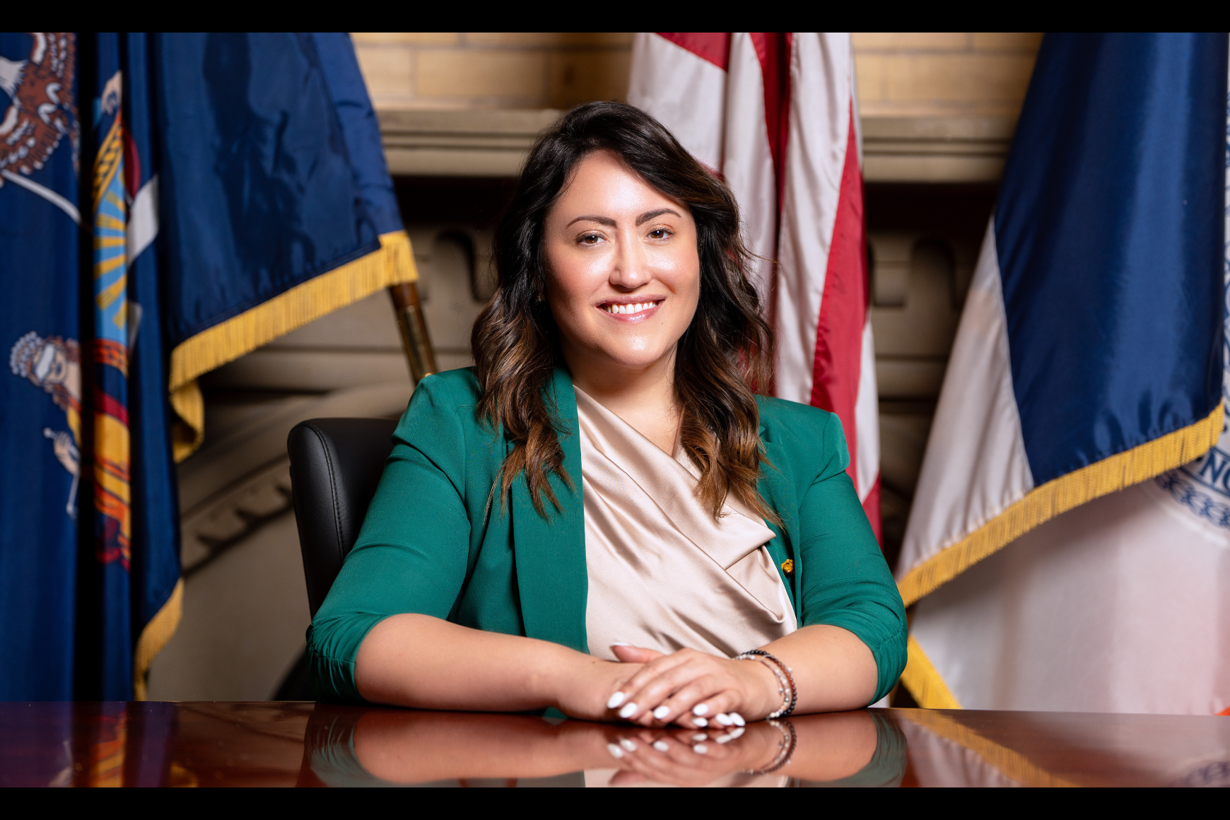 Woman in a green suit jacket sitting at a desk smiling in front of the American flag 